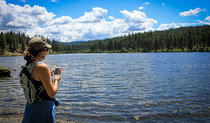woman on her smart phone standing next to a large scenic lake
