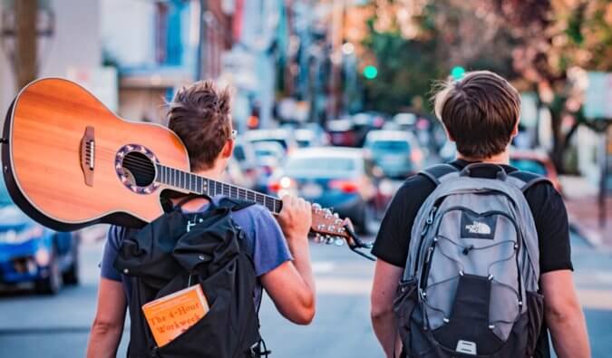 two backpackers walking down a street one is holding a guitar over his shoulder